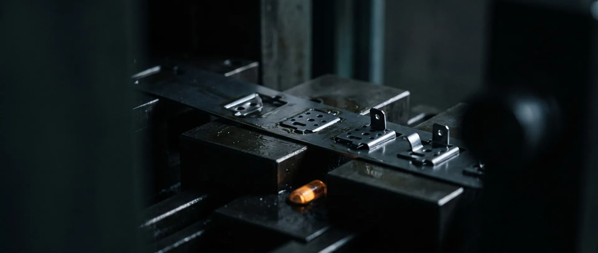 Macro of a progressive-die steel strip showing successive stations of a part being blanked, pierced and formed, oil sheen, dark press bed